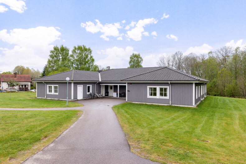 Boendet sett från uppfarten med gräsytor runt omkring. Byggnaden är grå i ett plan.
