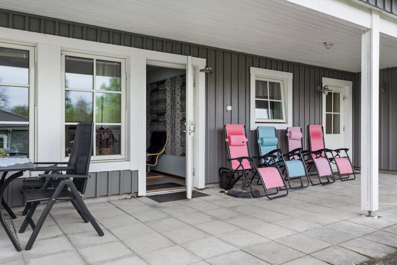 Öppen entrédörr vid en stenlagd uteplats med solstolar i olika färger.