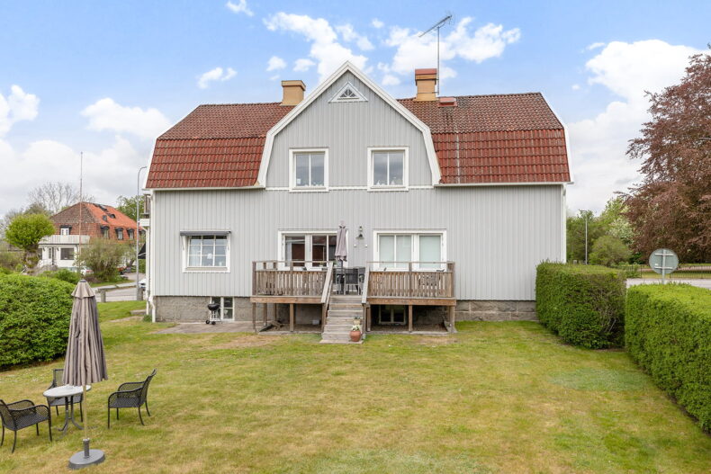 Gräsmatta, hög häck, altan, utemöbler och parasoll på gruppbostadens baksida.