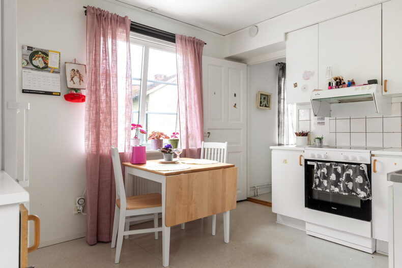 Kök i lägenhet med spis och skåp samt fällbart köksbord framför ljust fönster med rosa gardiner