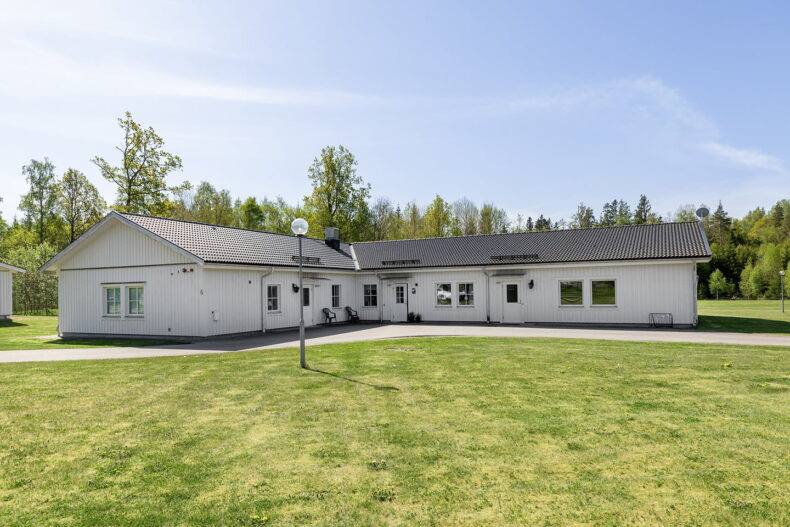 Boendets fasad, vit byggnad i ett plan omgivet av stora gräsytor.