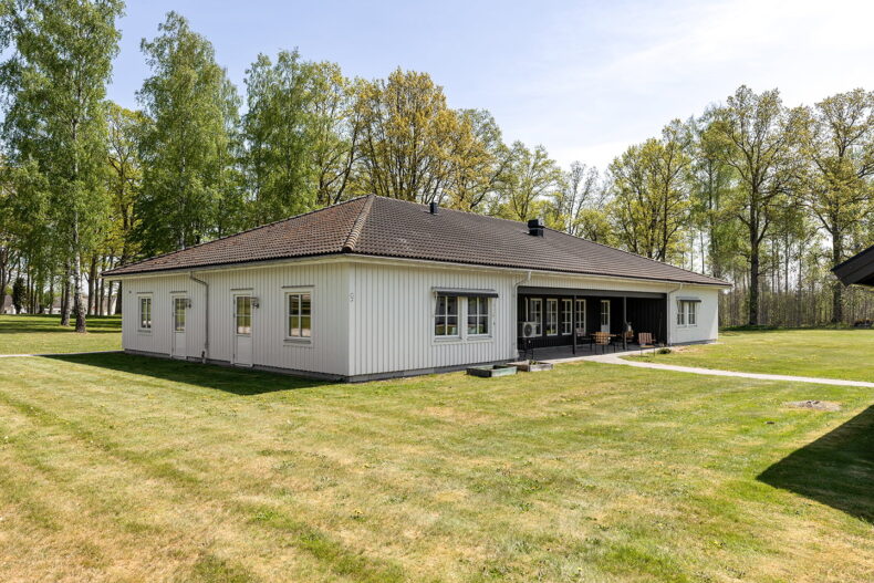 Boendets fasad, vit byggnad i ett plan, med stora gräsytor runtom och skog bakom