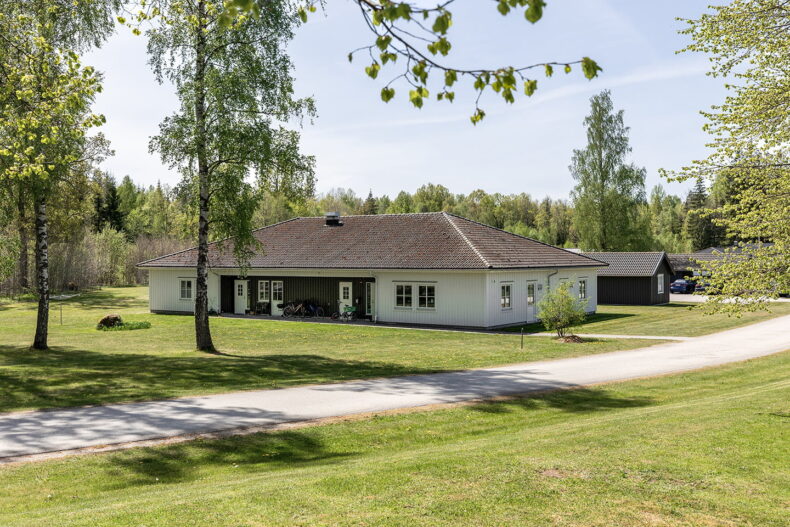 Boendets fasad, vit byggnad i ett plan, med stora gräsytor runtom och skog bakom