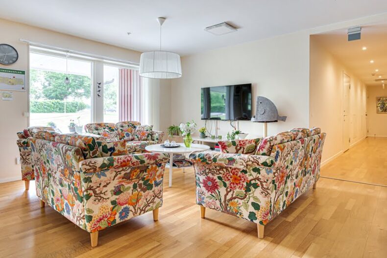 Ett gemensamt rum med blommiga soffor, TV och soffbord framför ett stort ljust fönster