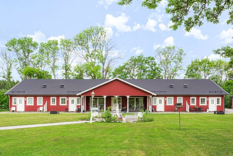 Boendet sett från andra sidan en stor gräsmatta. Byggnaden är röd med svart tak, i ett plan och mycket bred, med separata ingångar till lägenheterna.