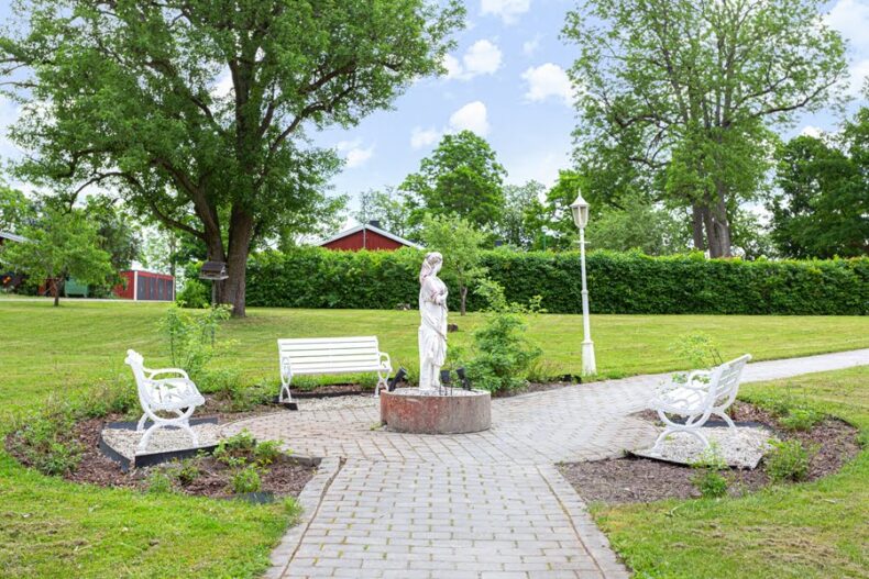 En stenlagd yta på gräsmattan med parkbänkar, rosenbuskar och en staty