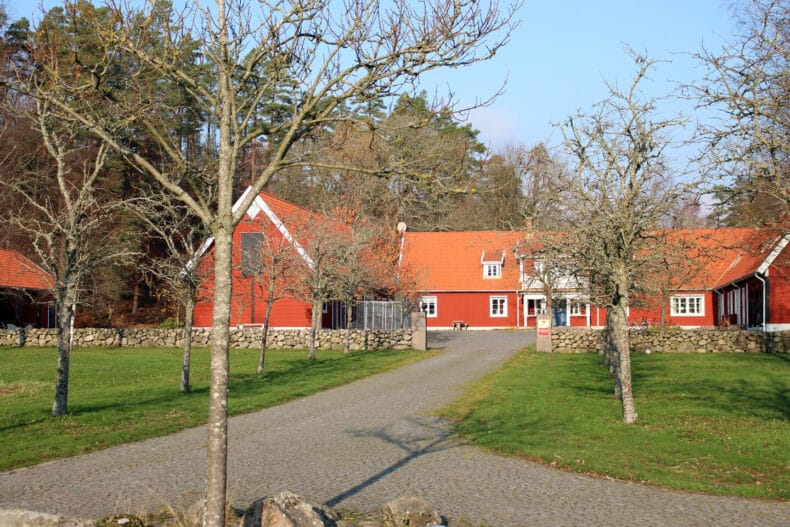 Boendet sett från uppfarten. Tre röda byggnader med två våningar och gräsyta med träd framför.
