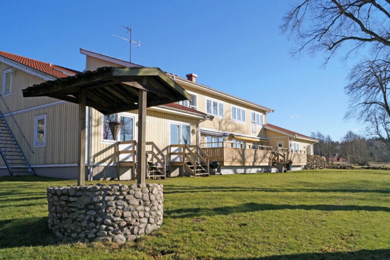 Boendets ljusgula fasad med altan och gräsmatta och brunn framför