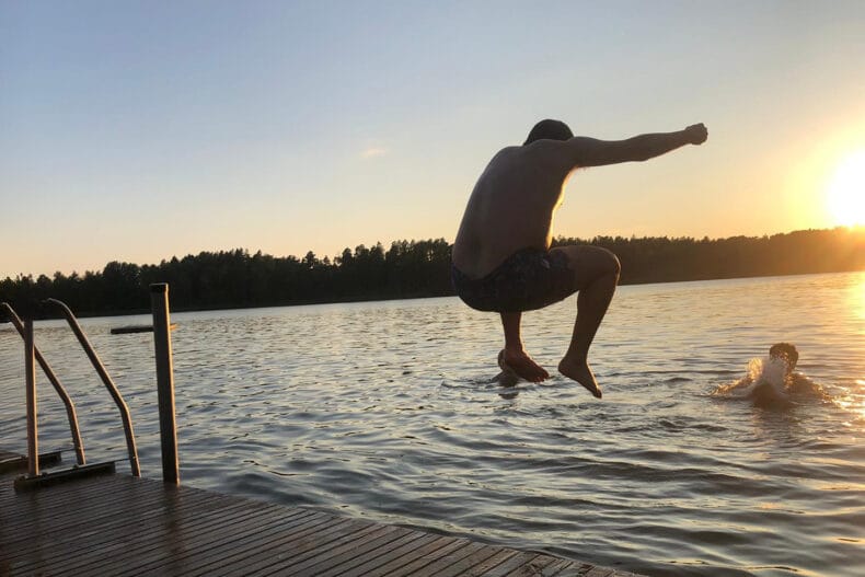 ”En person hoppar från en brygga ner i en sjö i solnedgången, medan en annan person redan badar i vattnet. Skog syns i bakgrunden och kvällssolen speglar sig i sjön.
